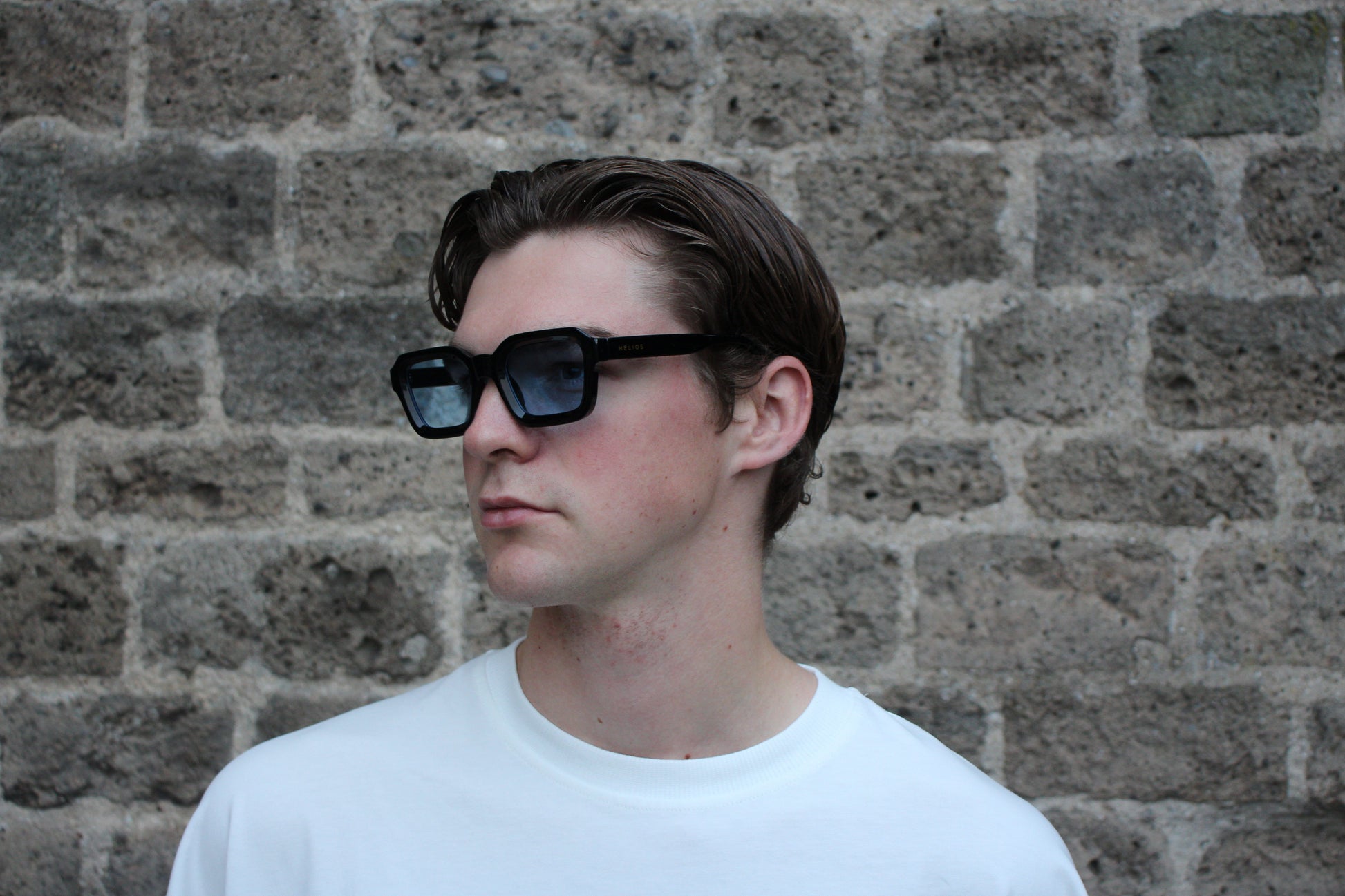 Person wearing sunglasses and a white shirt against a brick wall