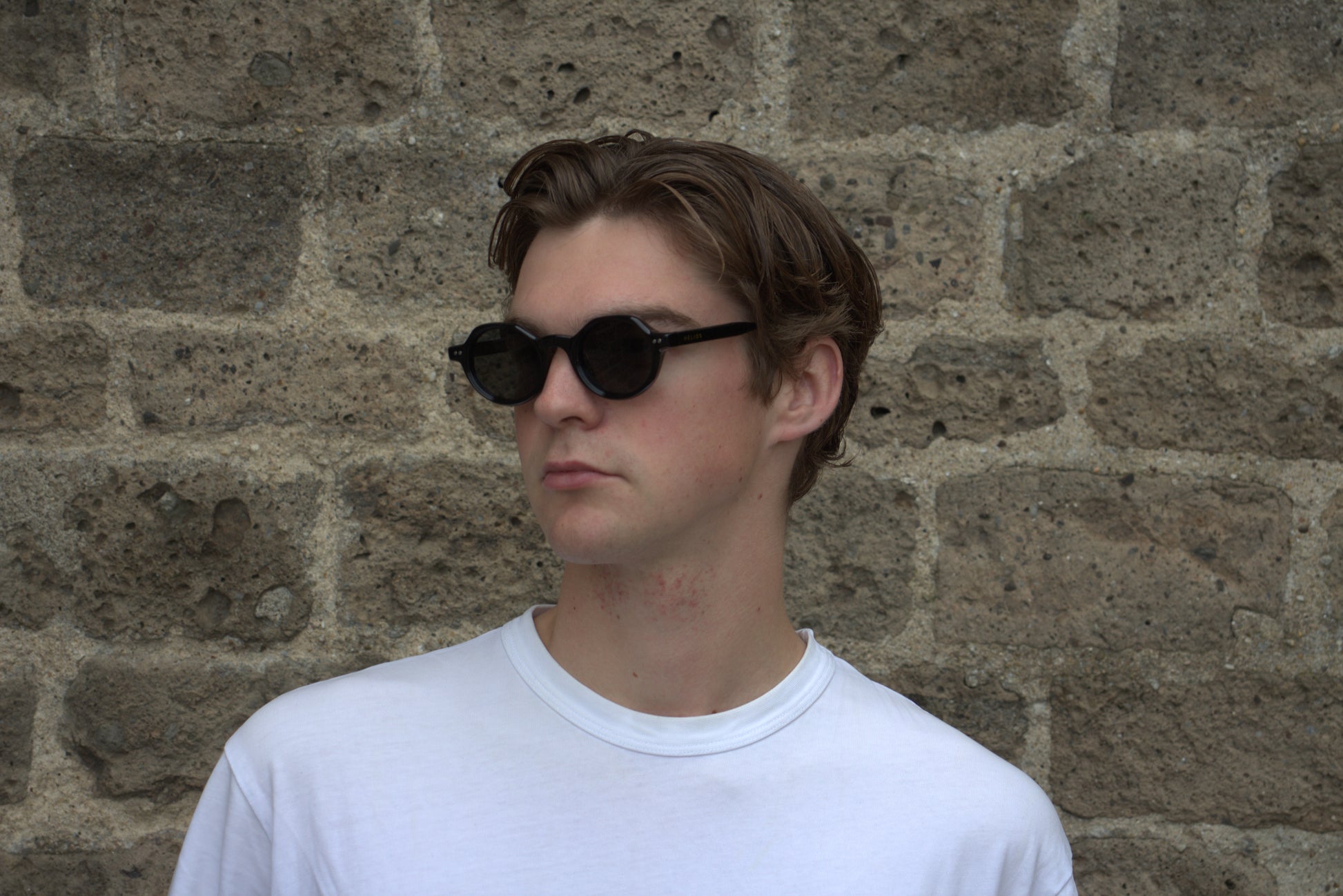 Person wearing sunglasses and a white shirt against a stone wall
