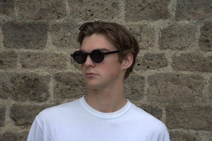 Person wearing sunglasses and a white shirt against a stone wall
